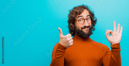 young bearded crazy man feeling happy, amazed, satisfied and surprised, showing okay and thumbs up gestures, smiling against flat color wall photo