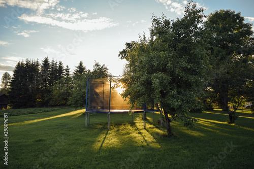 Open children's jump trampoline in the sun on green grass in the yard of finnish house with trees, summer evening