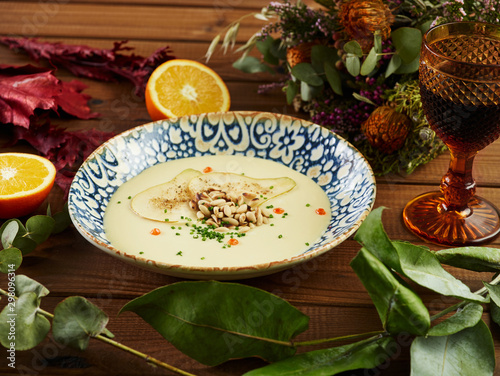 Bowl of delectable smoked leek vichyssoise with fresh orange and glass of wine placed on wooden tabletop near twigs of various plants