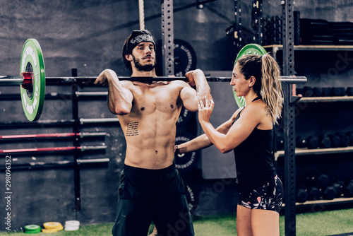 Strong coach in sportswear helping athletic client and doing barbell workout in modern gym