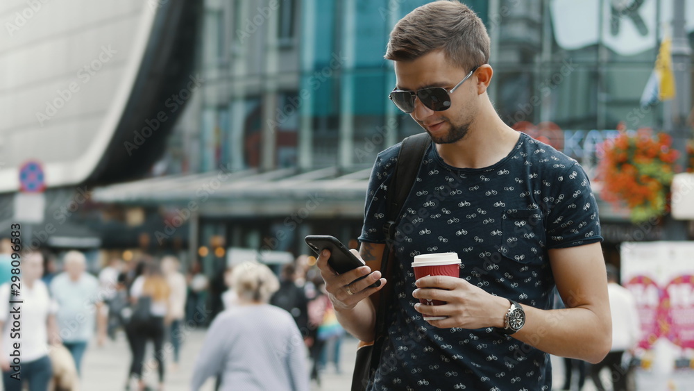 Modern man with a cup of coffee use mobile phone on crowded city street, wearing sunglasses, people and shopping center on background Modern man with a cup of coffee use mobile phone on crowded city street, wearing sunglasses, people and shopping center on background