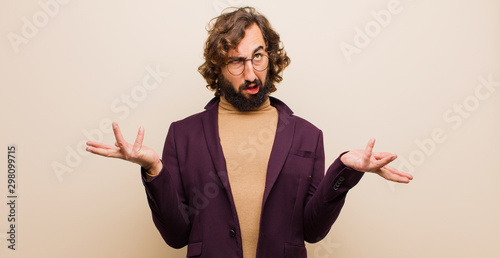 young bearded crazy man shrugging with a dumb, crazy, confused, puzzled expression, feeling annoyed and clueless against flat color wall photo
