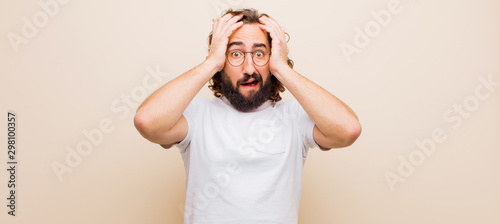 young bearded crazy man feeling horrified and shocked, raising hands to head and panicking at a mistake against flat color wall