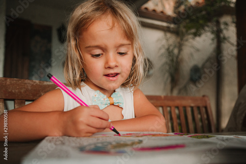 Focused little girl leaning on table and coloring pictures at book with marker pen on blurred background of room at home