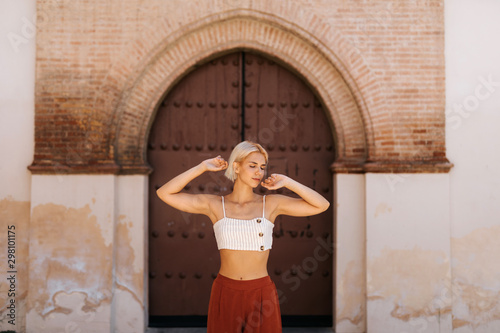 Attractive young woman in stylish outfit folding arms and closing eyes while standing against ancient building with shabby gate on street of old town