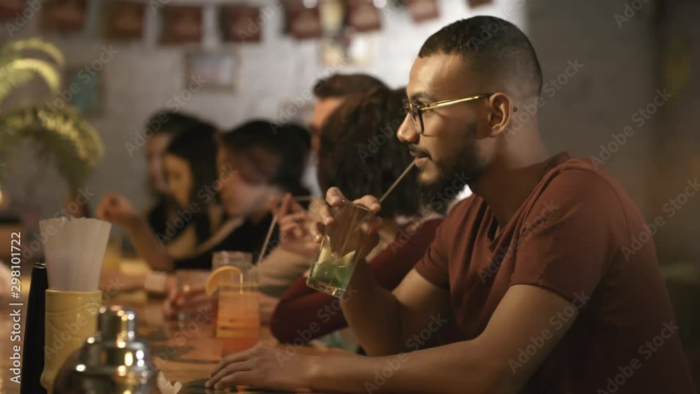 Side view medium shot of young bearded black man in glasses drinking cocktail at bar counter and talking to bartender, then turning head to camera Side view medium shot of young bearded black man in glasses drinking cocktail at bar counter and talking to bartender, then turning head to camera