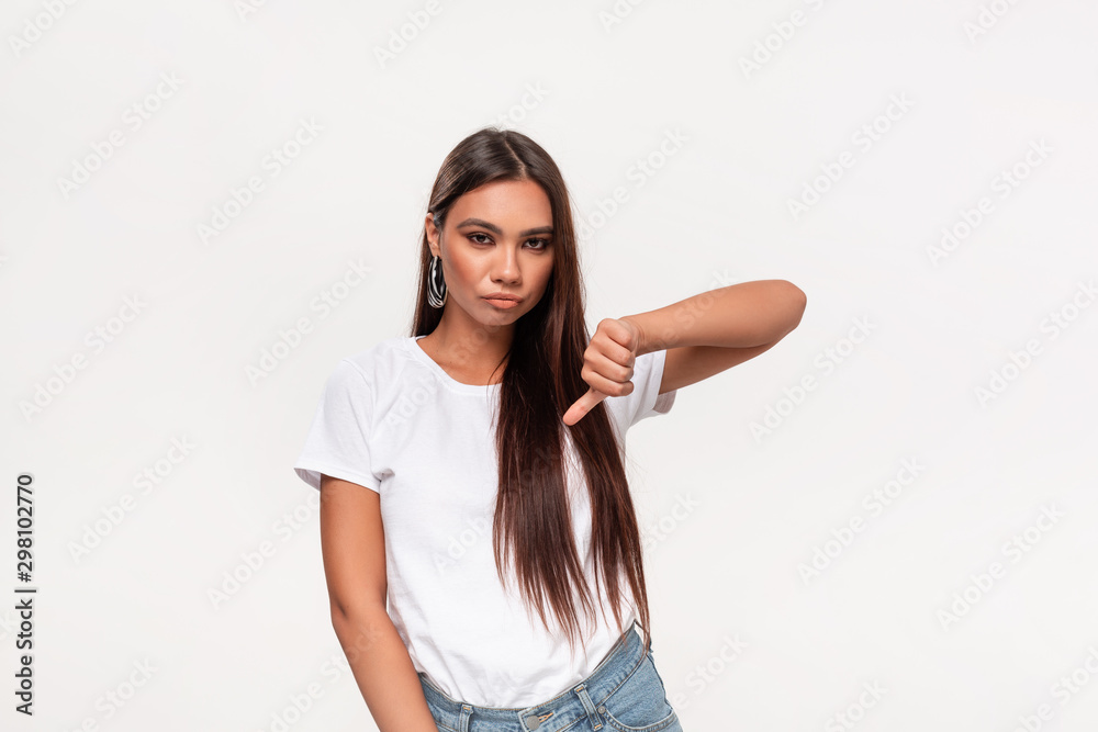 Upset pretty african-american teenager in a white t-shirt and blue jeans holding thumbs down isolated over white background. Concept of dislike. Upset pretty african-american teenager in a white t-shirt and blue jeans holding thumbs down isolated over white background. Concept of dislike.