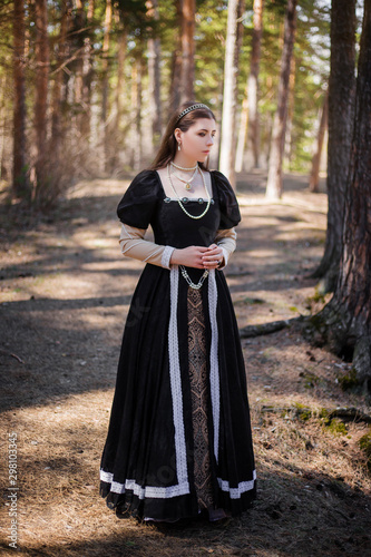 Young, beautiful girl in a black medieval dress walks through the gloomy pine forest.