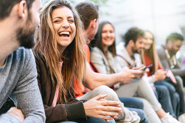 Group of trendy young people chatting together sitting on a bench outdoors. Students having fun together. Focus on a blonde woman smiling with open mouth