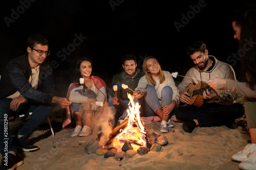 leisure and people concept - group of smiling friends sitting at camp fire on beach and roasting marshmallow at night
