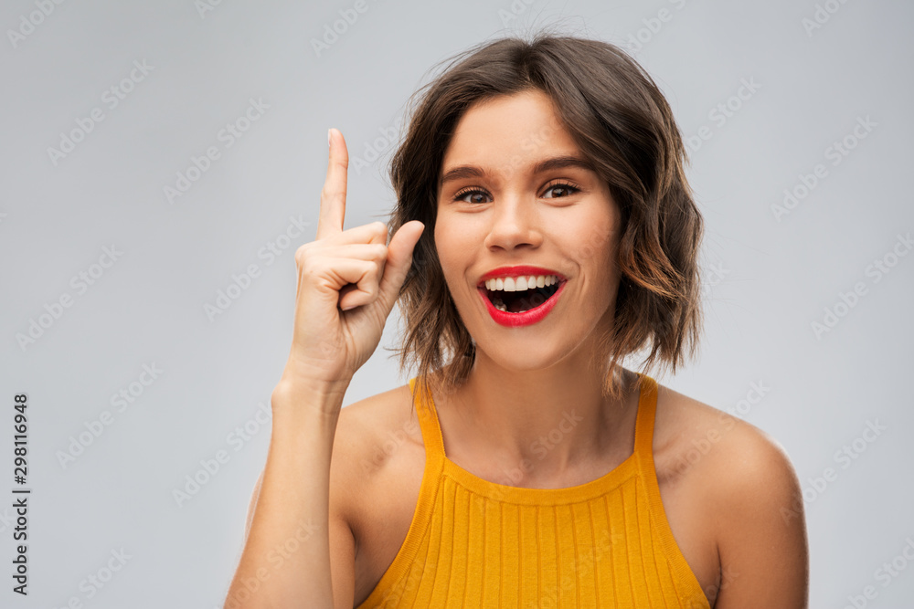 gesture, idea and people concept - happy smiling young woman in mustard yellow top pointing finger up over grey background gesture, idea and people concept - happy smiling young woman in mustard yellow top pointing finger up over grey background
