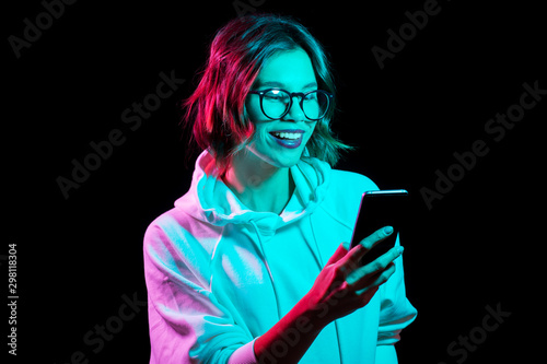 technology and people concept - happy young woman wearing hoodie and using smartphone in neon lights over black background