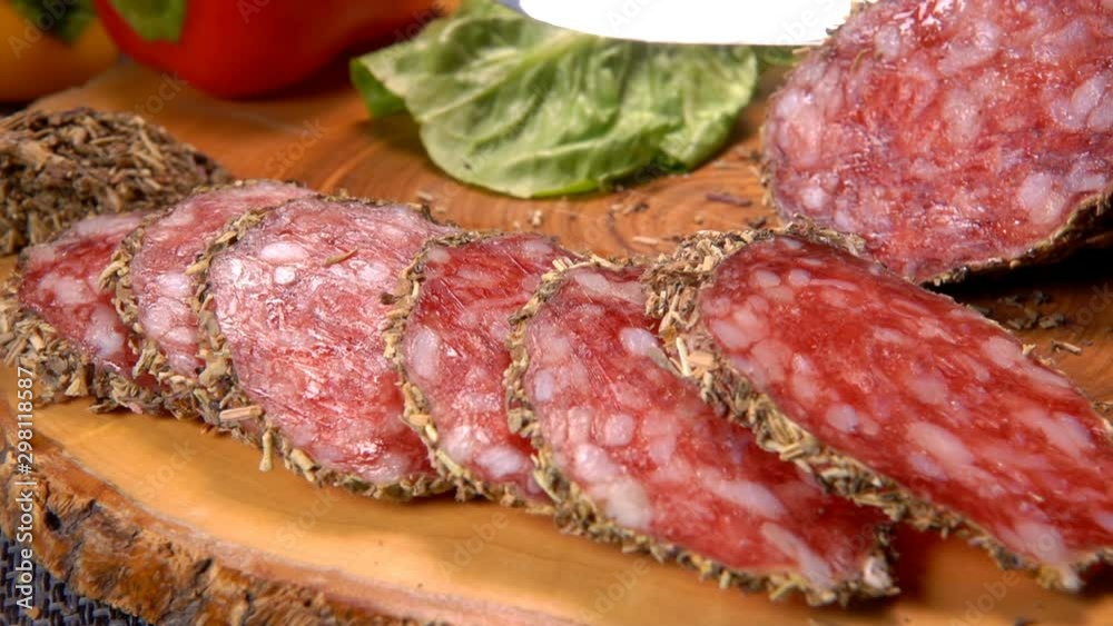 Sharp knife cut a slice of dried sausage in herbs on a wooden board on the background of greenery Sharp knife cut a slice of dried sausage in herbs on a wooden board on the background of greenery