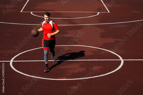 A bearded young basketball player in red sportswear is dribbling on a basketball outdoor court. Training and honing skills. Keeping the ball, effort, determination and achievement of the result. © Daniil