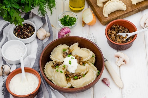 Boiled dumplings (vareniki) with potatoes and fried mushrooms with onions in a bowl with sour cream and green onions on a white wooden background.   Traditional Russian and Ukrainian dish.