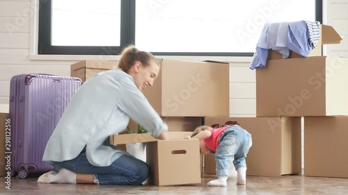 Little child looks in box. Background moving boxes and suit. Mother blonde woman wear blue shirt, jeans and white socks smile and look into box.