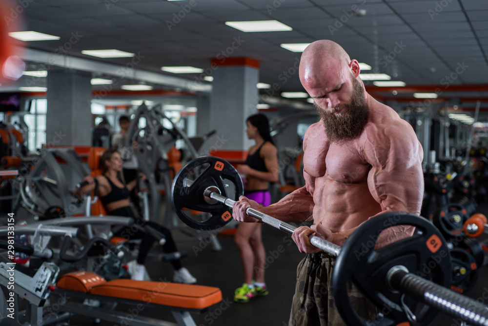 Obraz premium Bald man with a beard in the gym. Muscular bodybuilder guy doing barbell exercises. Strong man with a naked torso. The young athlete is preparing for weightlifting competitions.