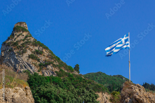 Greece flag waving in the blue sky with green fields at mountain peak background. Nature theme.