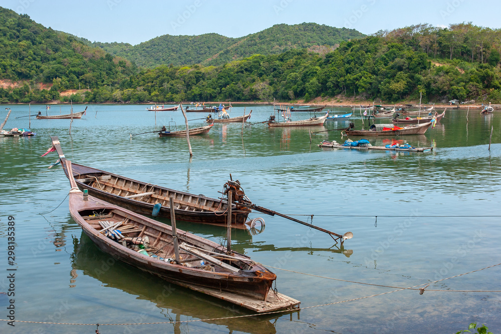 Many tethered long fishing boats with long propellers standing in the bay. On the shore hills overgrown with green trees.