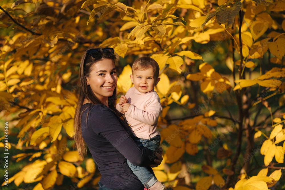 Portrait of beautiful family in the park. Mom and daughter togehter in autumn. Beautiful ten month baby on mother's hands. Background of yellow tree