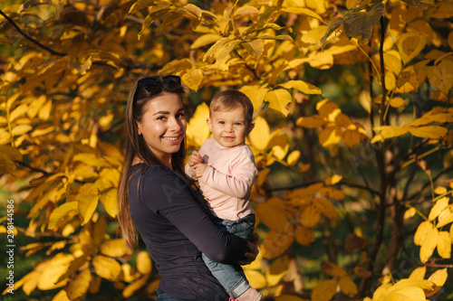 Portrait of beautiful family in the park. Mom and daughter togehter in autumn. Beautiful ten month baby on mother s hands. Background of yellow tree