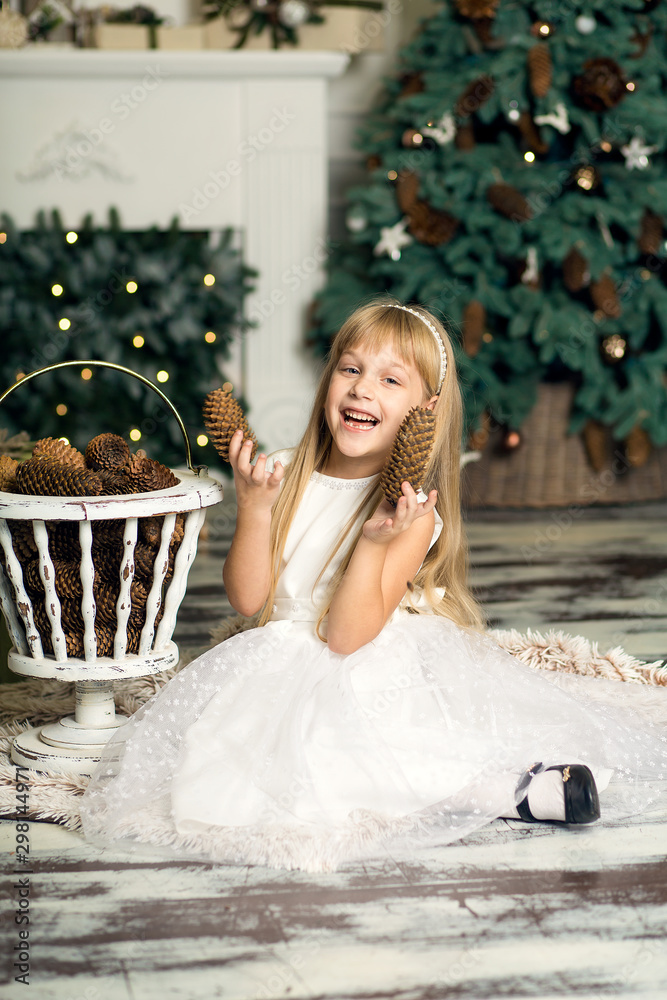 little girl in a white dress plays with pine cones in Christmas decorations against the background of the Christmas tree and fireplace. Merry Christmas and happy New Year