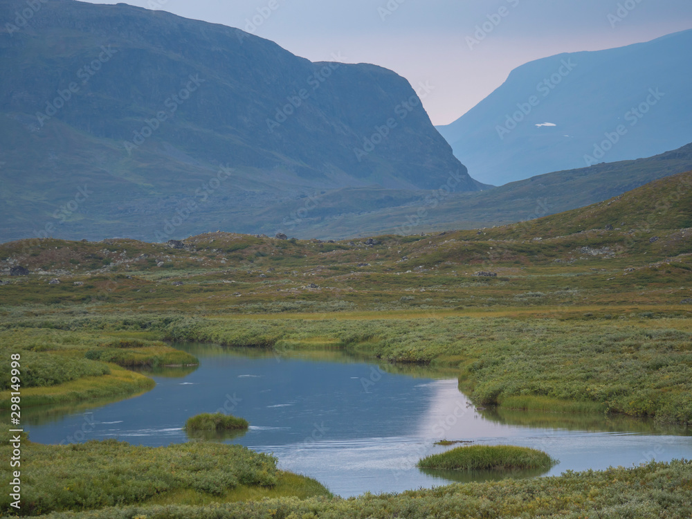 Beautiful wild Lapland nature landscape with blue glacial river, birch tree bushes, snow capped mountains and dramatic clouds. Northern Sweden summer at Kungsleden hiking trail.