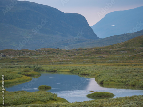 Beautiful wild Lapland nature landscape with blue glacial river  birch tree bushes  snow capped mountains and dramatic clouds. Northern Sweden summer at Kungsleden hiking trail.