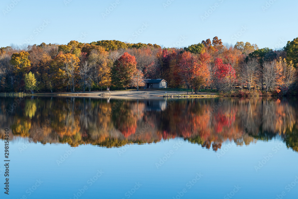 Empty beach shown from across the lake which also shows a reflection of the surrounding woods in the fall foliage colors