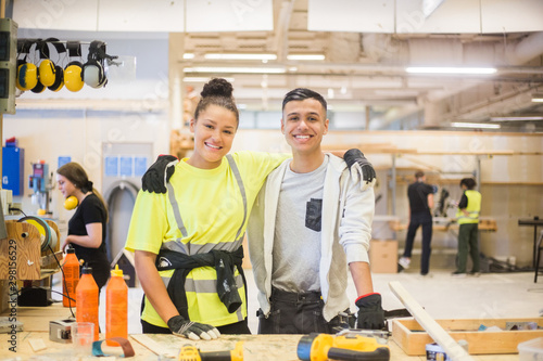 Portrait of smiling female trainee standing with arm around male coworker at workbench photo