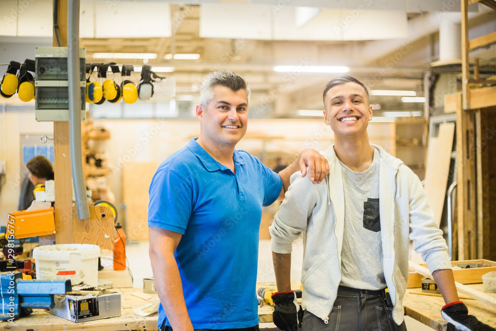 Portrait of smiling male instructor standing with hand on teenage trainee's shoulder at illuminated workshop Portrait of smiling male instructor standing with hand on teenage trainee's shoulder at illuminated workshop