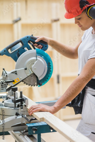 Young female trainee using electric saw for cutting plank at illuminated workshop photo