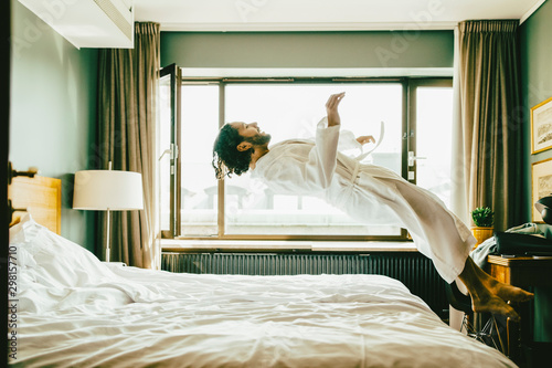 Playful man wearing robe jumping on bed at hotel