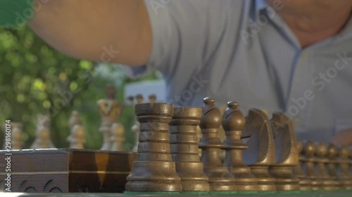 Old gray-haired man prepares the chess game outside in the garden at the table and puts the black figures on their position on the board.