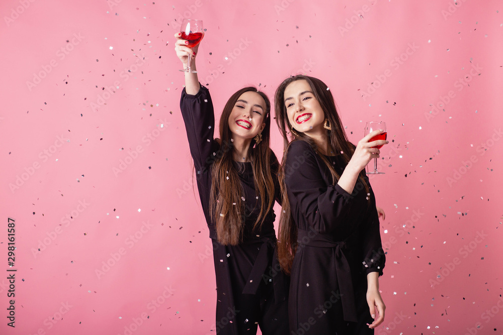 Two women celebrate the New Year party having fun laughing under the flying confetti and drinking wine.