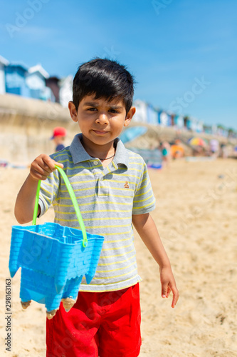 Indian kid 4-5 years in colorful wear, on a warm sandy beach, holding a blue beach bucket with beach huts in background photo