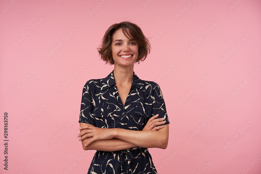 Indoor shot of pretty young brunette female with casual hairstyle wearing romantic dress, keeping hands folded and looking at camera with charming smile, isolated over pink background Indoor shot of pretty young brunette female with casual hairstyle wearing romantic dress, keeping hands folded and looking at camera with charming smile, isolated over pink background