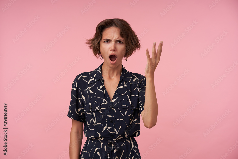 Studio shot of angry brunette female with casual hairstyle looking at camera with wide mouth opened and frowning face, posing over pink background with raised hand Studio shot of angry brunette female with casual hairstyle looking at camera with wide mouth opened and frowning face, posing over pink background with raised hand