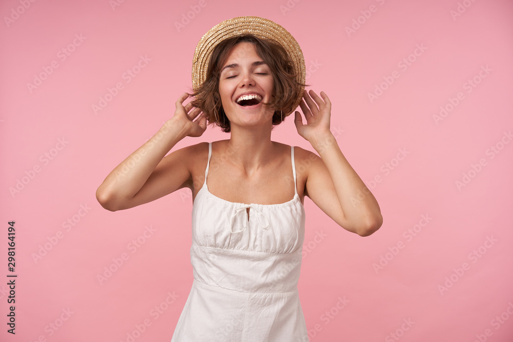 Indoor photo of pretty young female wearing white summer dress with straps and boater hat, keeping eyes closed while posing over pink background, being in high spirit Indoor photo of pretty young female wearing white summer dress with straps and boater hat, keeping eyes closed while posing over pink background, being in high spirit