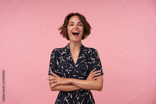 Indoor photo of joyful brunette female with casual hairstyle standing over pink background, keeping hands folded on her chest, looking at camera cheerfully with wide smile