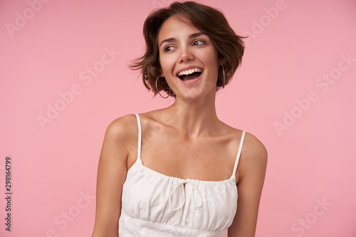 Studio shot of charming young female with casual hairstyle wearing white top while standing over pink background, looking aside cheerfully and smiling widely, being in nice mood
