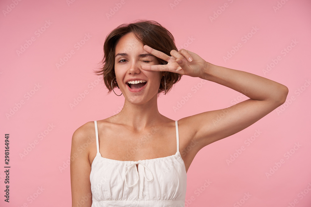 Cheerful young pretty lady with casual hairstyle wearing white top while posing over pink background, looking at camera joyfully with wide mouth opened and raising hand with victory sign