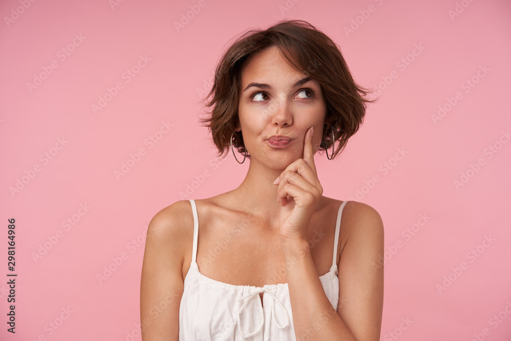 Indoor photo of lovely young brunette female with casual hairstyle wearing white top while standing over pink background, looking aside thoughtfully and rasing eyebrow