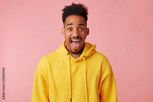 Indoor photo of young bearded dark skinned man looking at camera with wide eyes and mouth opened and wrinkling his forehead, wearing yellow sweatshirt while posing over pink background