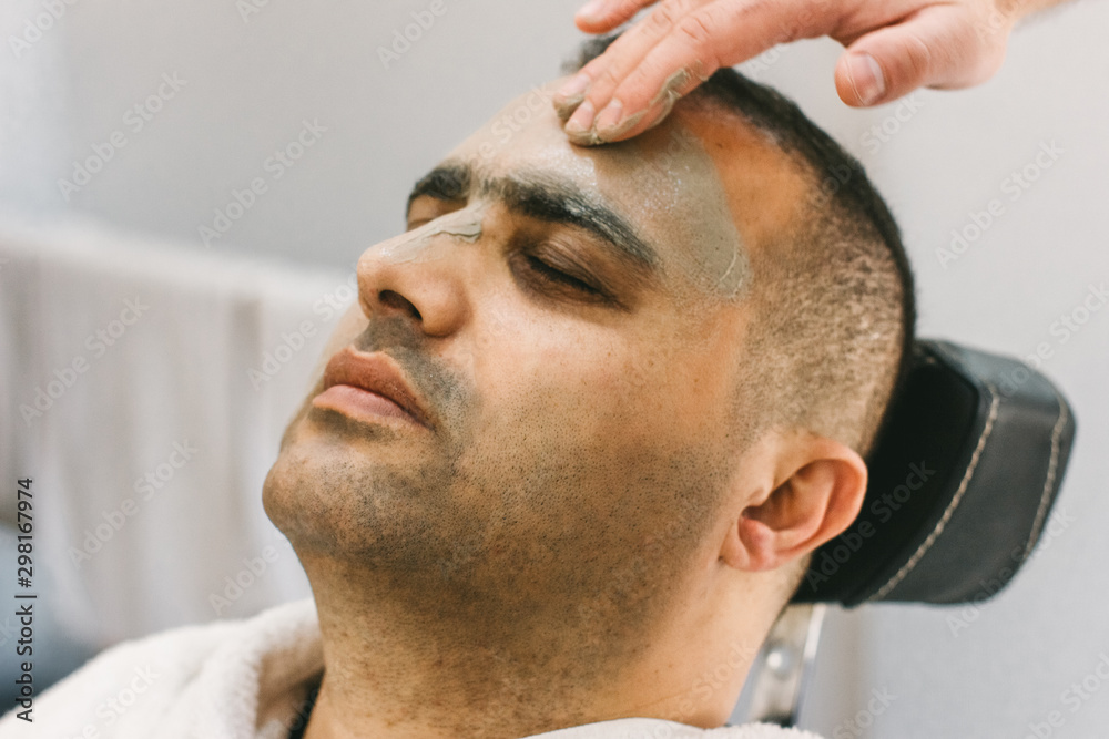 Obraz premium Male skin care in a beauty salon. Applying clay cleansing mask on a man's face