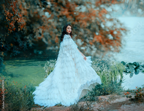 attractive brunette woman in luxurious fluttering waving outfit. A gentle snow queen walks. green lake. long Cloak with feathers. Spring Haze over water by river. Autumn foggy weather. Yellow trees photo