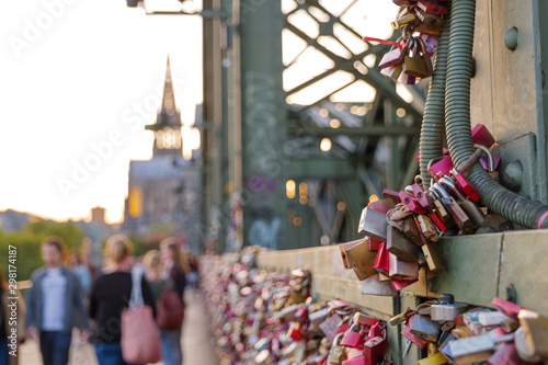 Close up view group of master key, and blurry crowd of people walk on pedestrian walkway beside steel truss arches structure of Hohenzollern Bridge and railway, and background of Cologne Cathedral.