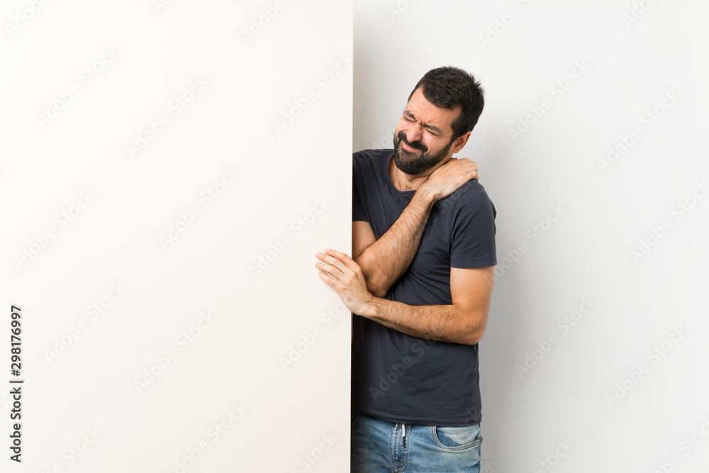 Young handsome man with beard holding a big empty placard suffering from pain in shoulder for having made an effort Young handsome man with beard holding a big empty placard suffering from pain in shoulder for having made an effort