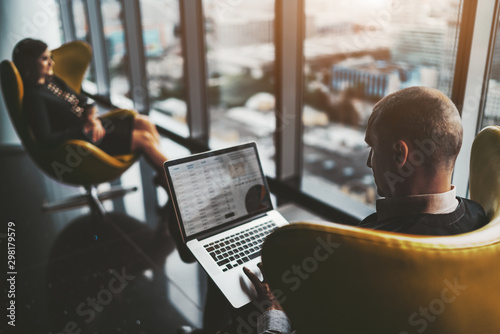 A business meeting near the window on the top floor of a luxurious office high-rise with cityscape outside: a man entrepreneur with the laptop and his female colleague aloof in a defocused background
