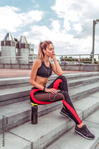 Girl athlete sits on stairs, rest after workout, summer day city. In hand, smartphone listens music headphones. Enjoys listens to podcast lecture. Active lifestyle fitness, fashionable stylish woman. © byswat